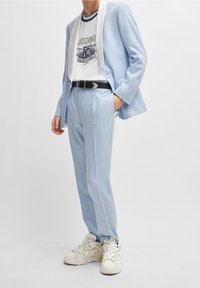 Costume sur mesure bleu clair avec des bordures blanches, associé à un t-shirt graphique blanc. Des baskets blanches avec des accents verts complètent le look.