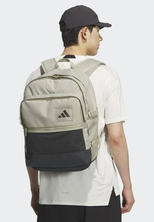 Beige and black backpack with multiple compartments, zippers, and a textured base. Features the Adidas logo on a front pocket.