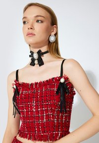 Punainen tweed-lyhyt toppi, jossa on mustia yksityiskohtia, fringed helmia ja helmiä muistuttavia yksityiskohtia, musta nauha-kaulaketju ja siihen sopivat korvakorut.