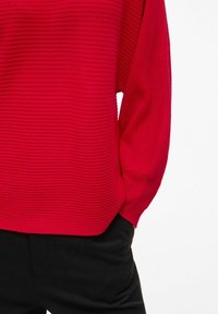 Pull à rayures rouges avec des manches longues ; texture douce et coupe ample. Associé à un pantalon en tissu noir pour un contraste.
