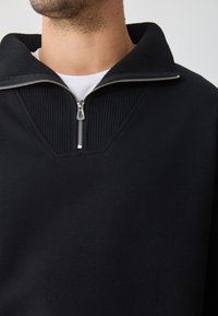 Pullover preto com colarinho canalé e design de meio zíper, feito de tecido suave, com uma silhueta minimalista e elegante.