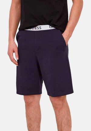 Homme portant un short bleu marine foncé avec la main dans la poche et un t-shirt noir, montrant une ceinture blanche avec le logo "GUESS".