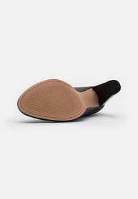 Chaussure noire avec un dessus en cuir lisse, un bout pointu et une semelle en caoutchouc beige présentant un motif de grip texturé.