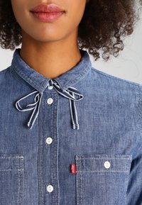 Camicetta in denim con collo a cravatta, bottoni bianchi e due tasche frontali. Include un'etichetta rossa con il logo Levi's.