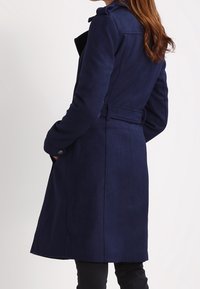 Marinblå, långärmad kappa med bälte i midjan, mjuk textur och skräddarsydd passform. Har en hög krage och sidofickor.