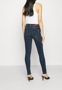 Jeans de denim azul oscuro, ajustados y de tiro medio, con bolsillos traseros y parche con logo. La modelo lleva una camiseta sin mangas blanca y sandalias negras con tacón.