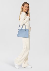 Borsa a tracolla in pelle azzurro chiaro con design strutturato, doppio manico e tracolla regolabile. Il modello indossa una maglietta color crema e pantaloni a gamba larga bianchi.