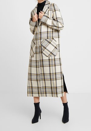 Personne portant un long manteau à carreaux crème avec de grandes poches et des bottines à talons noires, sur un fond uni.