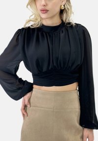 Schwarze, gekürzte Bluse mit langen, transparenten Ärmeln und gerafftem Oberteil. Kombiniert mit einem beigen, hoch taillierten Wildlederrock mit glatter Oberfläche.