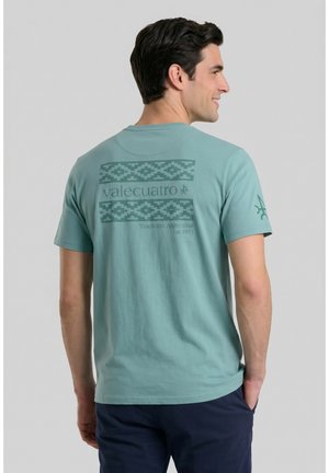 Hombre de espaldas con camiseta verde azulado con patrón geométrico y texto "valecuatro Tradición Argentina Est. 1971" en la espalda, pantalones oscuros.