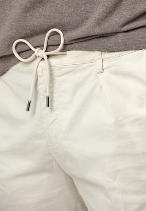 Helle beige Hosen aus Baumwolle mit flachem Schnitt, ausgestattet mit einem Kordelzugbund und Metallspitzen am Kordelzug. Glatte Textur.