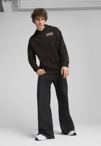 Sudadera negra con capucha y marca roja, bolsillo canguro y puños acanalados, combinada con unos jeans negros holgados y zapatillas blancas.