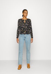 Blusa preta de manga longa com estampado floral e decote em forma de olho de fechadura, combinada com jeans azuis claros e botas de salto castanhas.