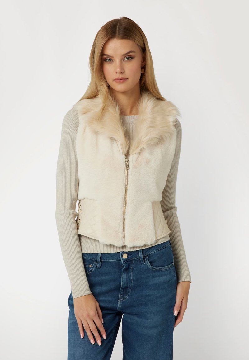 Kreminė dirbtinio kailio liemenė su užtrauktuku, turinti pūkuotą apykaklę ir deimantų rašto šonus. Dėvima ant pilkos raukšlėtos megztinės.