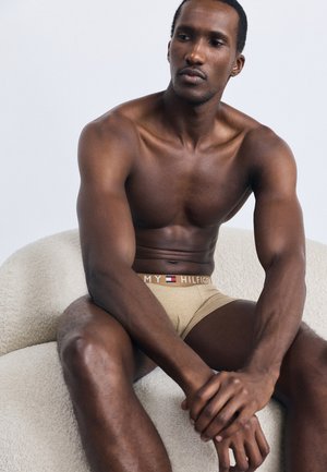 Beige pamut boxer sort márkás derékkal. A modellen izmos testalkat van, és egy textúrázott, világos színű kanapén ül.