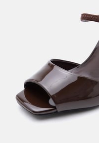 Σκούρο καφέ λουστρίνι σανδάλι με ανοιχτά δάχτυλα, τετράγωνη μύτη και καφέ λουράκι στον αστράγαλο ορατό στο πάνω δεξιά μέρος.