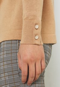 Manche de pull en tricot beige avec un poignet côtelé et deux accents de boutons. La main repose sur un tissu à motif tartan gris et noir.