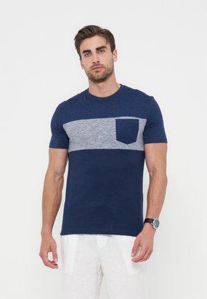 Άνδρας με μπλε σκούρο μπλουζάκι με γκρι οριζόντια ρίγα και τσέπη στο στήθος, σε συνδυασμό με λευκό παντελόνι και ρολόι χειρός, στέκεται μπροστά σε λευκό φόντο.