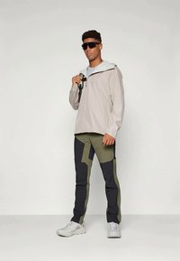 Lys grå regnjakke med hette, kombinert med grønne og marineblå fargeblokkede bukser. Modellen har solbriller og hvite joggesko.