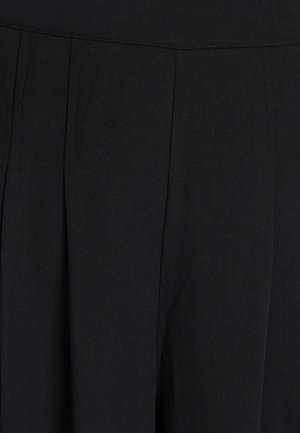 Melnas bikses, kas izgatavotas no struktūrēta auduma, ar krokotu priekšpusi un augstu jostasvietu. Minimālas detaļas un bez redzamiem aksesuāriem.