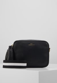 Czarna prostokątna skórzana torebka typu crossbody z złotym logo, bocznym frędzlem i odpinanym paskiem z czarno-białymi paskami na białej powierzchni.