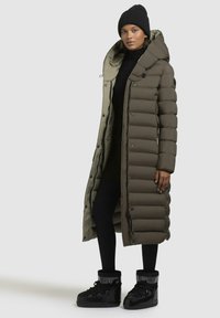 Long, puffy olive green coat with a hood, featuring large quilted sections and black accents. Worn with a black beanie and black high-tops.