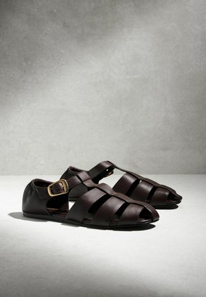 Mørkebrune lædersandaler med flere stropper, spænde ved anklen og flad sål, vist på en lysegrå overflade med bløde skygger.