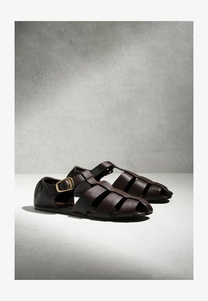 Mørkebrune lædersandaler med flere stropper, spænde ved anklen og flad sål, vist på en lysegrå overflade med bløde skygger.