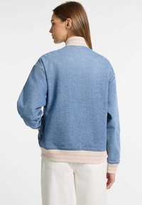 Denim bomber bunda v svetlom modrom odtieni, s rebrovými manžetami a spodným lemom a prúžkovaným detailom na golieri. Má uvoľnený strih a mäkkú textúru.