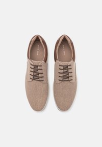 Zapatos de tela beige con acentos de cuero marrón, punta redonda, suela plana y cordones marrones. La etiqueta dice "PIER ONE" en la plantilla.