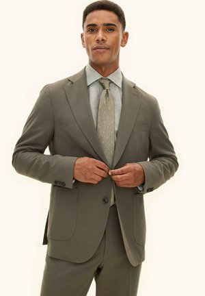 Uomo che allaccia la giacca di un completo verde oliva sopra una camicia verde chiaro con una cravatta a motivo, su sfondo semplice.