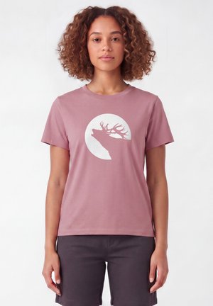 Vrouw met krullend haar die een mauve T-shirt draagt met een witte silhouet van een huilende hert in een cirkel en donkere shorts.