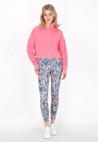 Rosa Zip-up Sweatshirt mit hohem Kragen, kombiniert mit bunten, abstrakt gemusterten Leggings. Das Modell trägt weiße Sneaker.
