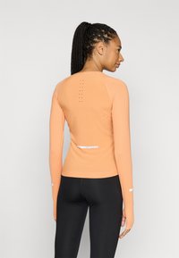 Ilgos rankovės sportinė palaidinė šviesiai oranžinės spalvos audinyje, su šoninėmis ir nugaros perforacijomis, atspindinčiomis detalėmis ir prigludusiu siluetu.