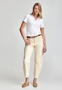 Camisa polo blanca con cuello, pantalones tapered color crema y mocasines color marrón claro. Diseño simple, textura de algodón y detalle de cinturón marrón.