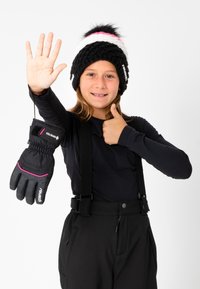 Bonnet en tricot noir avec des rayures blanches et roses, orné d'un pompon. Le modèle tient un gant noir avec des accents roses et fait un signe de pouce levé.