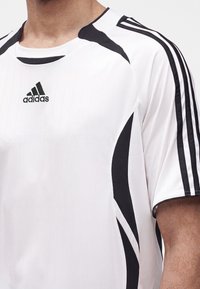 Λευκό αθλητικό μπλουζάκι Adidas με μαύρες ρίγες στους ώμους και στα πλαϊνά, με το λογότυπο της Adidas στο στήθος, φορεμένο από ένα άτομο.