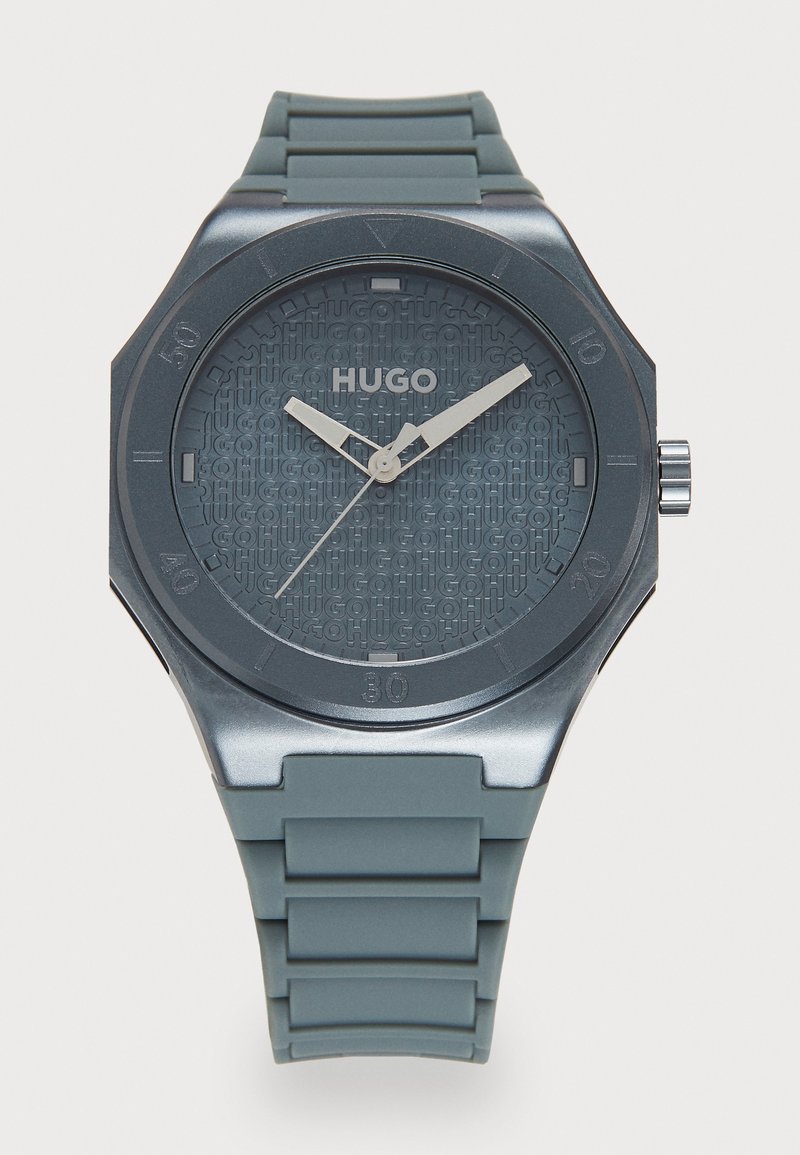 Montre Hugo avec un bracelet en caoutchouc gris foncé, un boîtier octogonal et un cadran texturé présentant un logo répété. Des aiguilles argentées indiquent l'heure.