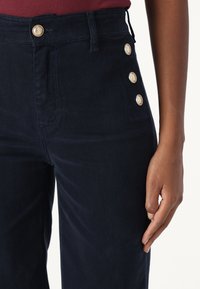 Primer plano de pantalones azul oscuro con botones dorados en la cinturilla y en el lateral, combinados con una blusa color marón, mostrando una mano descansando sobre el muslo.