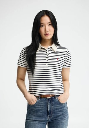 Femme aux longs cheveux noirs portant un polo rayé blanc et bleu marine et un jean bleu, debout les mains dans les poches devant un fond uni.