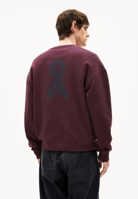 Felpa color bordeaux con un grande logo testurizzato blu scuro sul retro, colletto rotondo e polsini a costine. Abbinata a jeans neri.