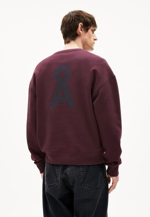 Bordeaux hoodie met een groot, getextureerd logo in donkerblauw op de achterkant, ronde halslijn en geribbelde manchetten. Gecombineerd met zwarte jeans.