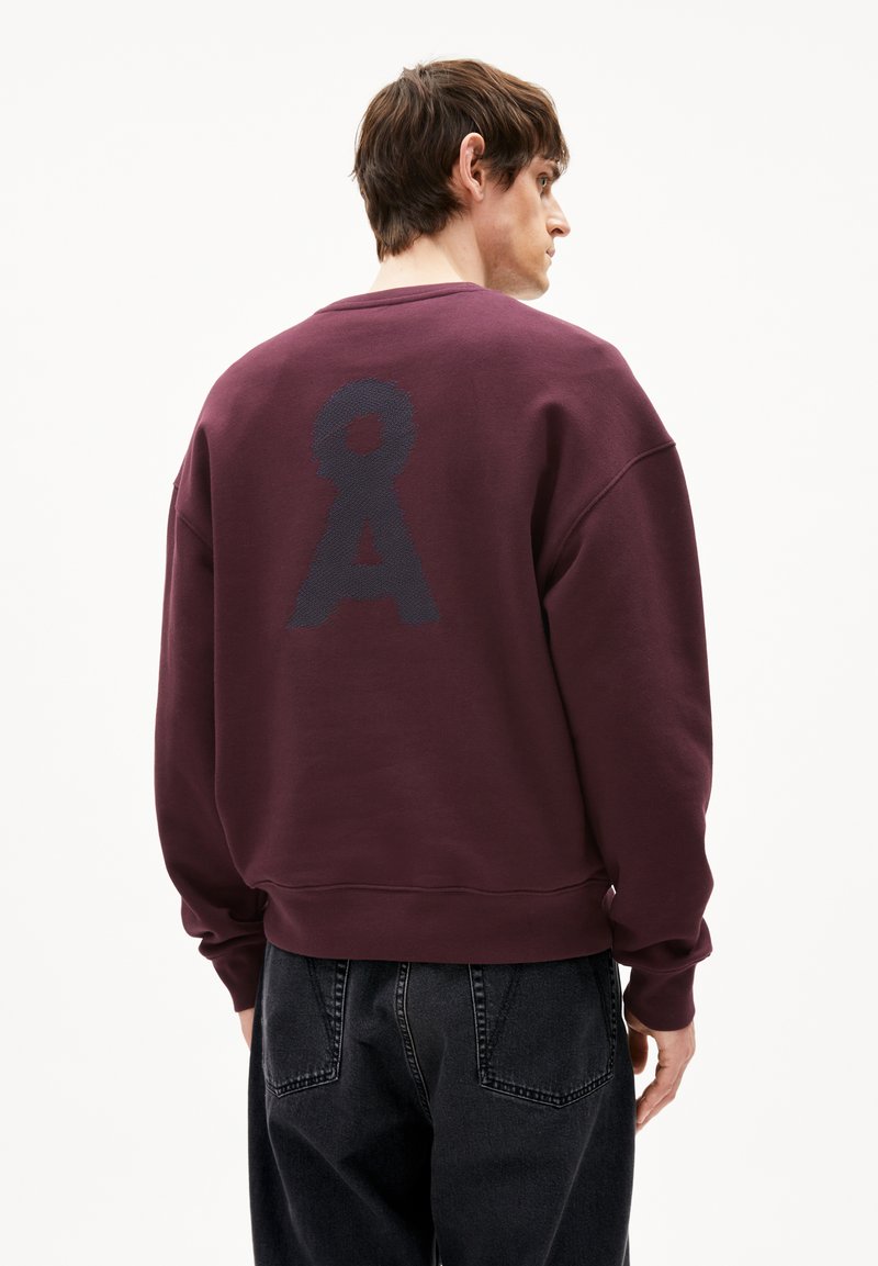 Felpa color bordeaux con un grande logo testurizzato blu scuro sul retro, colletto rotondo e polsini a costine. Abbinata a jeans neri.