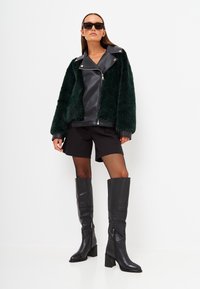 Groene faux fur jas met zwarte leren accenten, strakke shorts, zwarte visnet panty's en knielange zwarte laarzen met hakken.