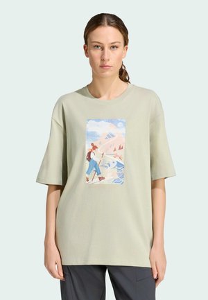 Vrouw draagt oversized beige t-shirt met kleurrijke hiking-geïnspireerde afbeelding van persoon met rugzak en wandelstokken nabij water en bergen.