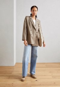 Dobbeltbrystet beige blazere, lyseblå vidde jeans, beige platform-sko og en skuldertaske med lang rem. Glat tekstur.