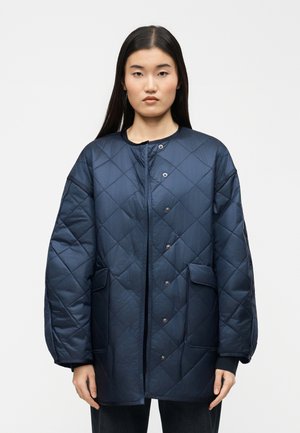 Femme aux longs cheveux foncés portant une veste matelassée bleu marine avec boutons-pression et grandes poches avant, debout devant un fond uni.