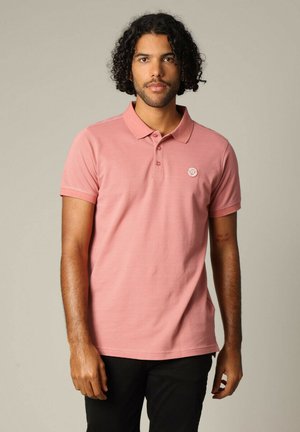 Polo rose avec un tissu texturé, des manches courtes et une patte de boutonnage à deux boutons. Présente un logo sur la poitrine gauche. Porte avec un pantalon noir.