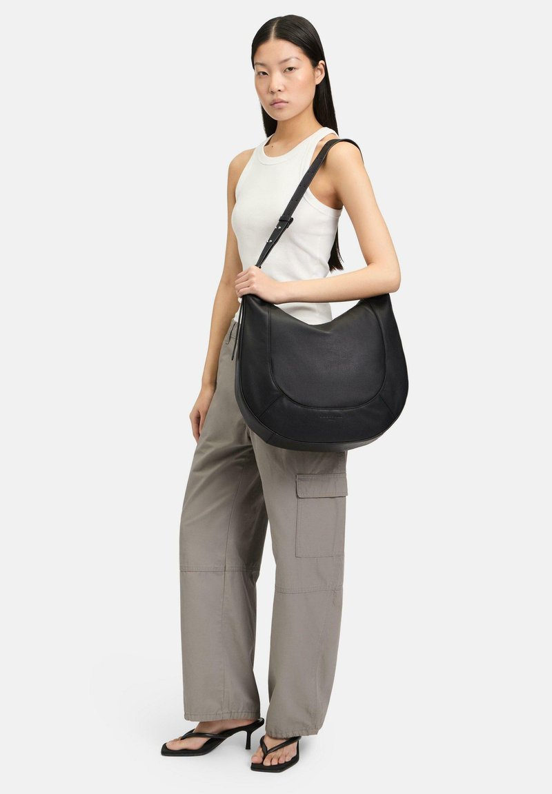 Sac à main en cuir noir avec un design incurvé, une surface texturée et une bandoulière ajustable. Le modèle porte un haut blanc ajusté et un pantalon cargo.