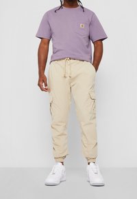 Muž oblečený v levandulovém tričku Carhartt, béžových cargo jogger kalhotách a bílých teniskách, stojící na pozadí z čistě bílé barvy.